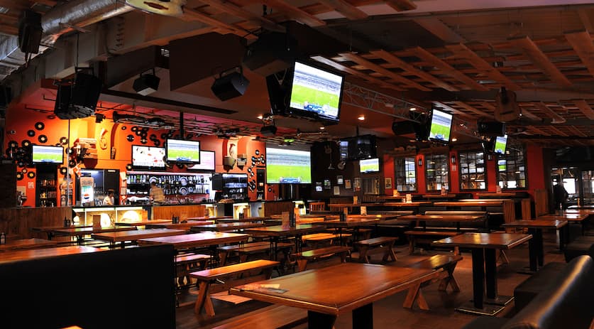 Spacious sports bar layout with wooden dining tables, bench seating, a bar counter, and multiple TV screens mounted throughout