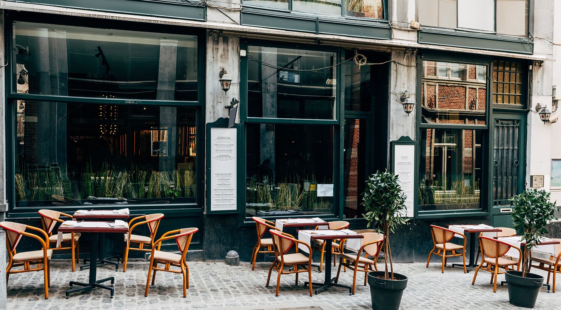 A restaurant storefront with dark green facade, large windows, outdoor seating with wicker chairs and tables on a cobblestone sidewalk.