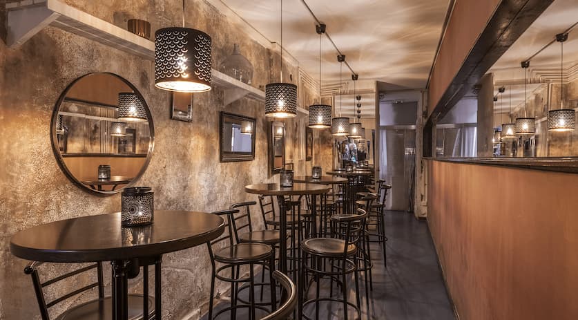 Bar interior with round high-top tables and metal bar stools along a counter under decorative pendant lighting